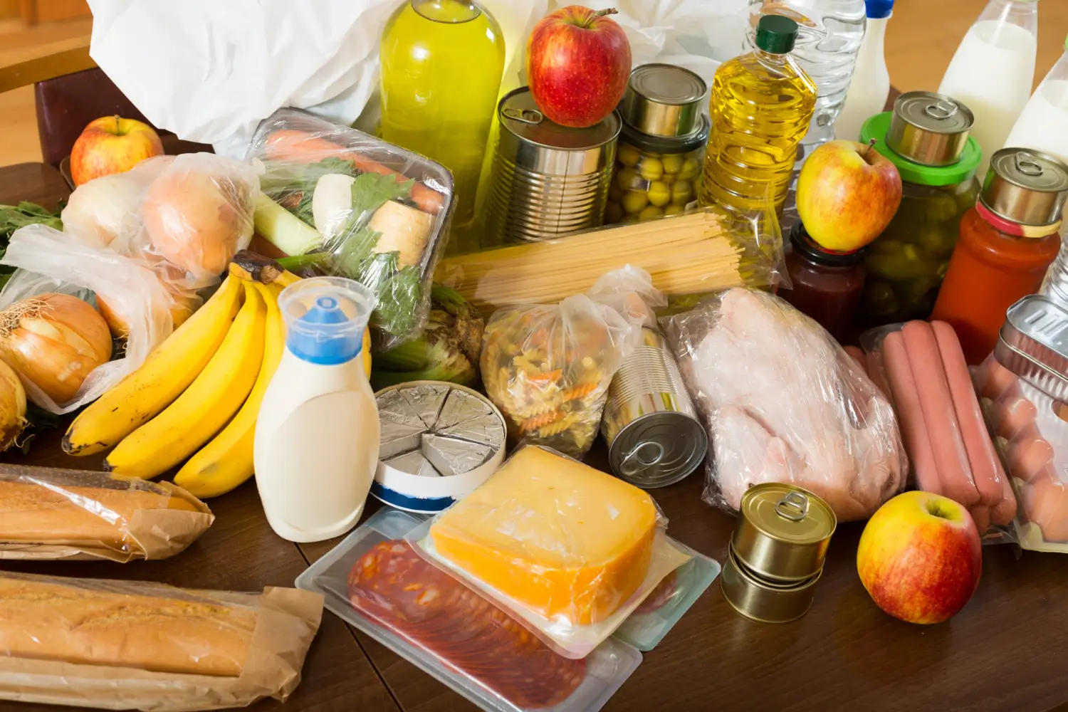 Mesa com compras de supermercado: frutas, latas, garrafas de óleo, leite, frios e pães